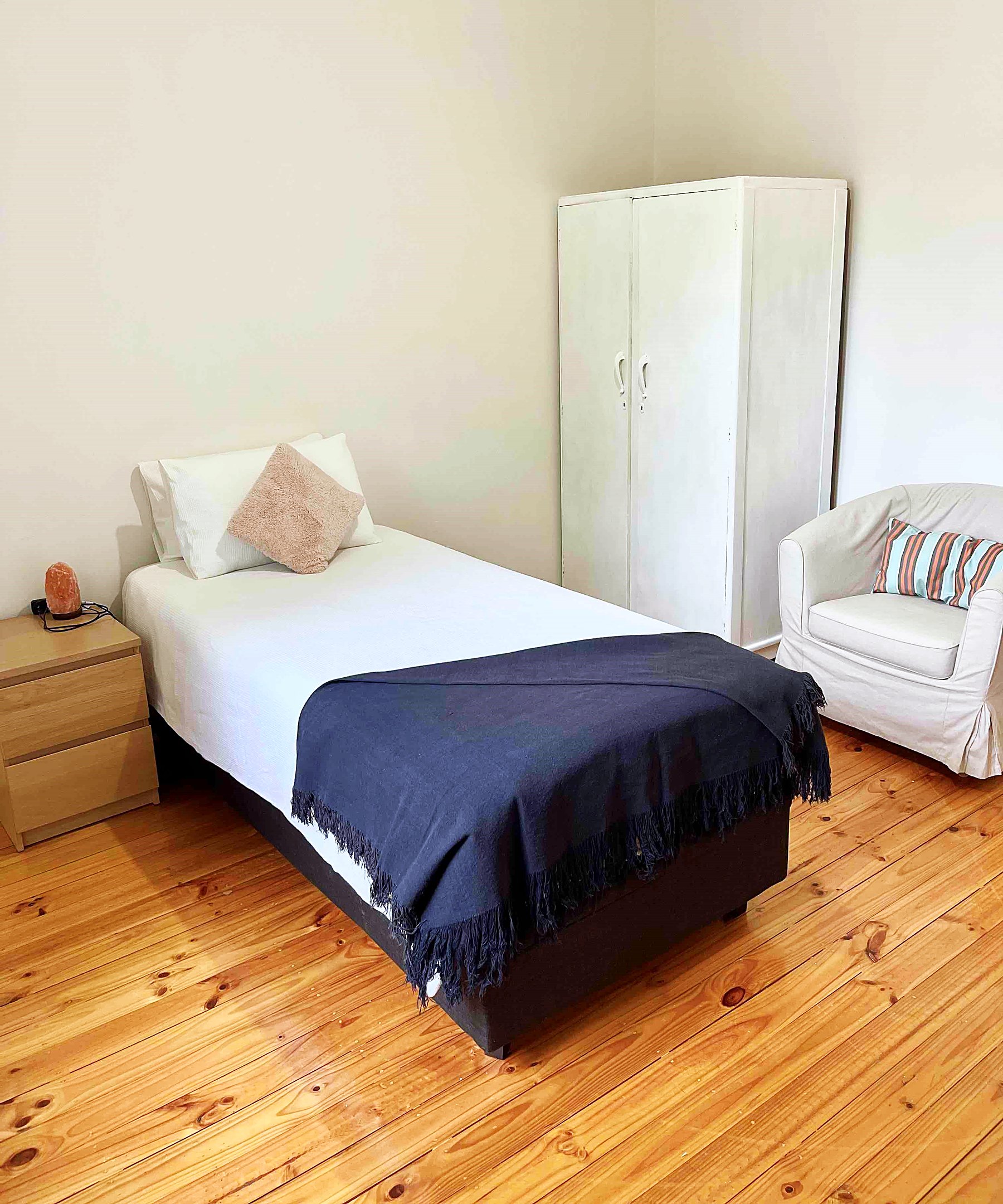 A furnished single bedroom with a wardrobe, armchair, bedside table and lamp